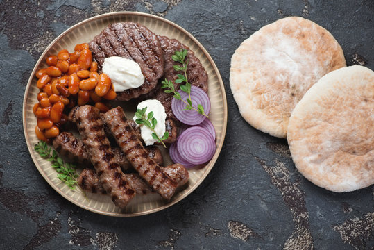 Above view of a round beige tray with grilled balkan cevapcici, pljeskavicas, kajmak cheese, prebranac and pitas over brown stone background