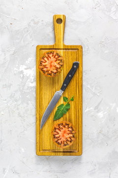 Slices raw fresh tomato and kitchen knife on wooden cutting board. Concept of green house life style and products of subsistence farming, flat lay on light gray concrete surface, copy space
