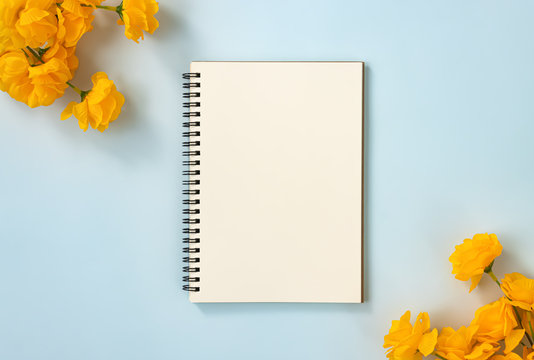 Spiral Notebook or Spring Notebook in Unlined Type and Orange Flowers at Bottom Right and Top Left Corner on Blue Pastel Minimalist Background. Spiral Notebook Mockup on Center Frame