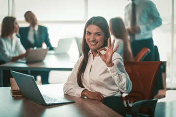 businesswoman shows ok sign working in open space office. Happy Caucasian woman with laptop showing alright gesture against her coworkers on blurred background. Tinted image.