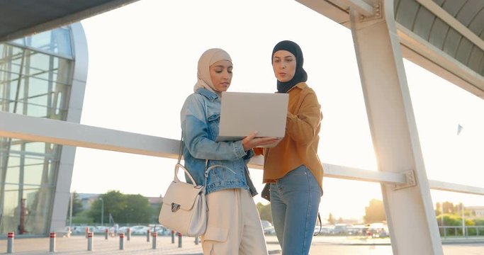 Two Caucasian young females friends in traditional Arabic headscarves standing at street and using laptop computer. Muslims friendly beautiful women alking and discussing something on screen.