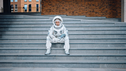 Sad Man in Spacesuit is Sitting on Concrete Stairs. Astronaut is Feeling Down and Looks at the Camera. Emotionally Depressed Spaceman in White Futuristic Suit with Technological Panel on His Hand.