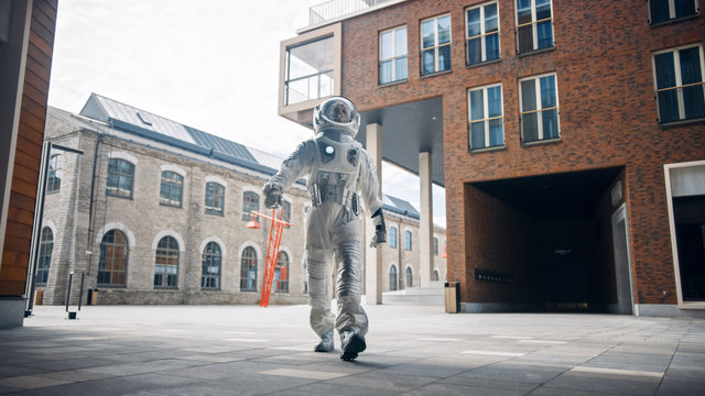 Low Angle Shot Of A Confident Handsome Astronaut Is Walking Towards The Camera In A Neighbourhood With Modern Scandinavian Buildings. Man In Futuristic Suit With Technological Panel On His Hand.