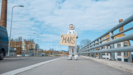 Man in Spacesuit is Standing at the Edge of a Road and Holding a Sign with Mars Written on it....