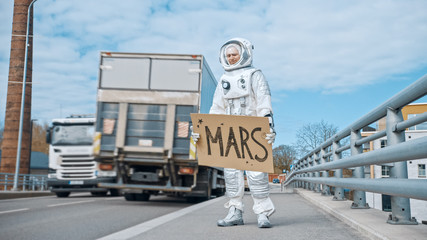 Man in Spacesuit is Standing at the Edge of a Road and Holding a Sign with Mars Written on it....