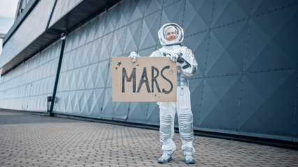 Handsome Man in Spacesuit is Standing Outside Next to an Industrial Metal Wall and Holding a Carboard "Mars" Sign. Spaceman in Futuristic Suit with Technological Panel on His Hand.