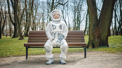 Sad Man in Spacesuit is Sitting on a Bench in a Park. Miserable Astronaut Looks in the Distance. Emotionally Depressed Spaceman in White Futuristic Suit with Technological Panel on His Hand. © Gorodenkoff
