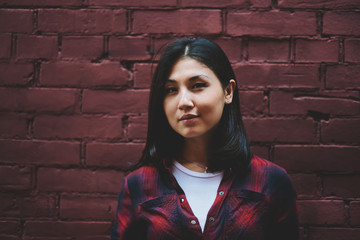 Close up portrait of young trendy brunette hipster girl standing near wall and posing, confident attractive asian female teenager 20s in fashionable stylish apparel looking in camera outdoors