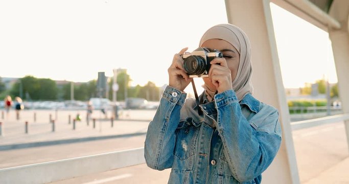 Muslim young beautiful stylish woman in traditional headscarf taking photos on vintage photo camera at street. Female in Arabic hijab making pictures while standing outdoor in city.