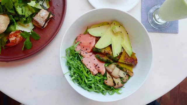 Waiter Hand Puts Salmon Salad with Avocado, Zucchini, Mushrooms and Green Leaves on White Table in Restaurant. Top View. Poke Bowl with Raw Fish and Vegetables. Smoothie. Fresh Healthy Delicious Food.