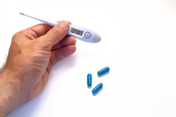 hand with a digital thermometer showing fever values and blue pills over white background