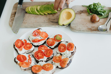 Process of making toasts with avocado on modern white kitchen. Home cooking concept. Hand putting avocado slices on sandwich made of whole grain bread,  tomato, and cheese.