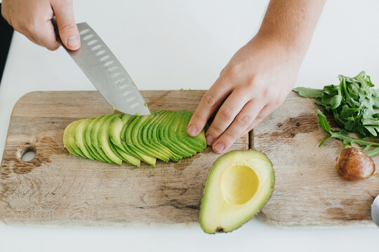 Home Cooking Concept. Person Cutting Avocado For Sandwich On Modern White Kitchen. Process Of Making Healthy Toasts With Avocado, Tomato, Arugula, Cheese And Whole Grain Bread.