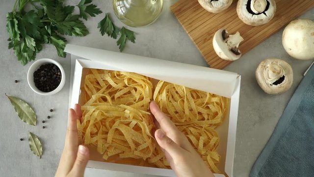 Top view of female hands pack box with with cooked raw Fettuccine pasta on background of mushrooms and olive oil and greenery.