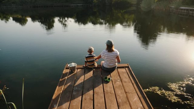 Happy mother teaches her little son to fish while sitting on a wooden pier by the lake on a sunny summer day