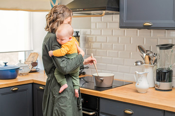 mom with baby in her arms in the kitchen kitchen preparing food - obrazy, fototapety, plakaty