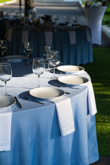 Served tables for a banquet or wedding in the summer outdoors. Tables with a blue tablecloth, white empty plates, cutlery and wine glasses. Served round table for guests