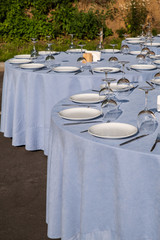 Served tables for a banquet or wedding in the summer outdoors. Tables with a blue tablecloth, white empty plates, cutlery and wine glasses. Served round table for guests