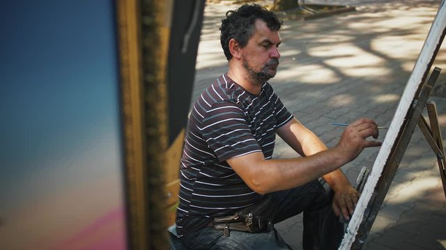 Male artist drawing picture on canvas using oil paints while sitting on an alley of city park