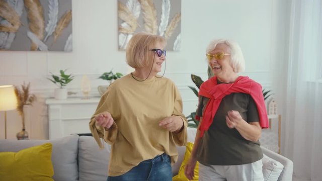 Couple of happy beautiful old fashion grandmas dancing out together in stylish apartment living room. Friendship. Two grandmothers having party. Slow motion.