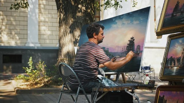 Male artist working at picture, creating marine landscape with sailing ships while sitting in park