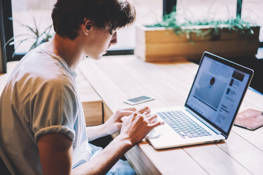 Skilled male IT professional creating program code for creating new application on laptop device during remote work in cafeteria, young millennial man dressed in casual look reading news via app