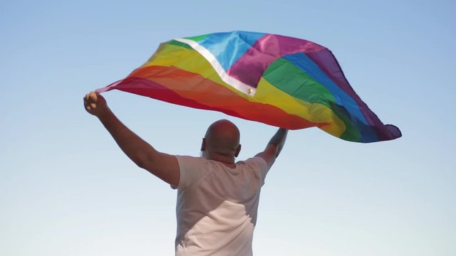 Bold man with rainbow gay flag while parade on background of blue sky. LGBTQI, Pride Event, LBGT Pride Month, Gay Pride Symbol
