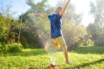 Funny little boy playing with garden sprinkler in sunny backyard. Preschooler child laughing,...