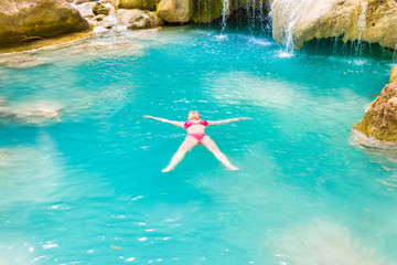 Red haired young woman in pink bikini swimsuit swims in emerald blue water of tropical lake. Erawan National park, Kanchanaburi, Thailand