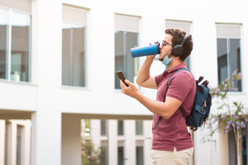 Attractive student drinking while the mobile phone with the surgical mask on his neck.