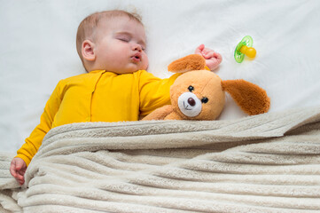 Portrait of a sleeping baby in bed under a blanket with a toy and pacifier nearby