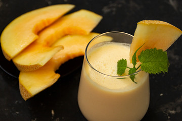 Melon milk shake on a black background. Ripe sliced melon is lying nearby. The smoothie glass is...