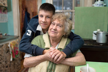 Teenager grandson hugs his grandmother in the kitchen at home. Happy family relationships, love, caring for an old woman. Poor, ordinary people.
