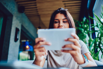 Concentrated young hipster girl playing video games online on tablet spending time in cafe interior, female student enjoying watching interesting movie on portable pc during leisure in coffee shop