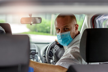 Photo of handsome young man wearing face mask sitting in a car,view from rear seat. Driver taking to a passenger on seat back wearing protective medical mask. One man with protective mask riding a car