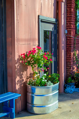 A painted wooden bench and a stock tank full of flowers and plants make a colorful rustic decoration outside a doorway to an old warehouse.