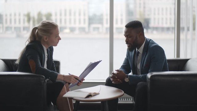 Young HR woman interviews afro-american male candidate for job sitting on couch in conference room