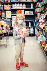 A new normal. Caucasian blonde girl in sanitary face mask shopping at toy store. Child wearing protective mask against coronavirus. Safety, health protection during covid-19 quarantine.