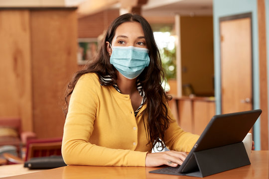 Portrait Of Young Ethnic Woman Wearing Yellow Sweater With Black And White Striped Blouse And Face Mask, Sitting At Bar In Kitchen Of Downtown Loft With IPad 