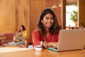 Portrait of young ethnic woman wearing orange blouse sitting at bar in kitchen of downtown loft using laptop computer