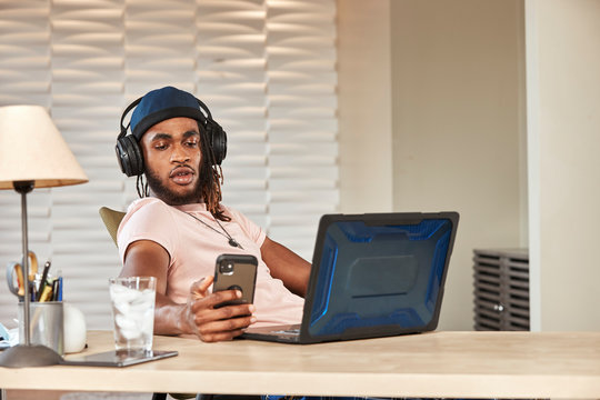 portrait young ethnic man sitting at desk with laptop computer wearing large over ear headphones looking at smartphone 