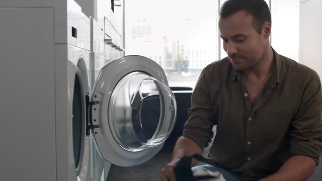 Young Man Loading Clothes Into Washing Machine In Kitchen