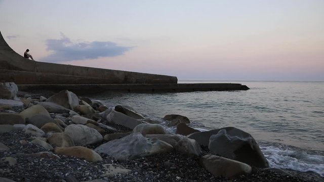 A young man with curly hair in a cap uses his laptop while sitting on a rocky beach on a concrete breakwater in the evening during sunset. Perhaps he is a freelancer and he works remotely.