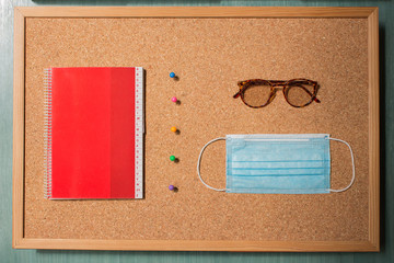 A red notebook with alphabetical index separated by a row of colored pins from vintage eyeglasses and a surgical mask with a corkboard in the background. Back to school concept.