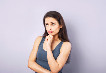 Fototapeta premium Frustrated grimacing brunette woman thinking and looking up in grey t-shirt with folded arms on purple background with empty copy space. Closeup