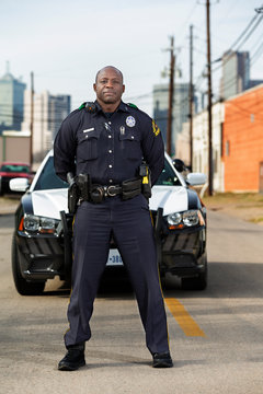 Portrait of Police officer Standing in street in front of squad car looking towards camera with arms behind his back 