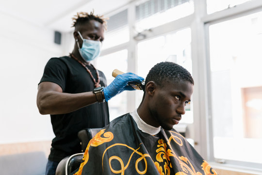Barber wearing gloves and mask cutting young man's hair with razor in salon