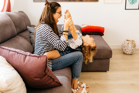 Mother with mouth open holding playful daughter's hands while sitting with her on sofa at home