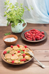 Pancake cereals on the fork. Red ripe fresh raspberries in a brown plate and a bunch of Melissa in a mug, yogurt on a dark wooden table near the window. Delicious Breakfast