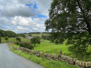 Looking across a country road, with dry stone walls, fields, trees and farm buildings on a cloudy day near, Denholme, Bradford, UK